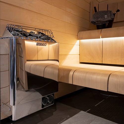 Harvia KIP30B electric sauna heater stainless steel with sauna stones beside wooden bench and warm lighting