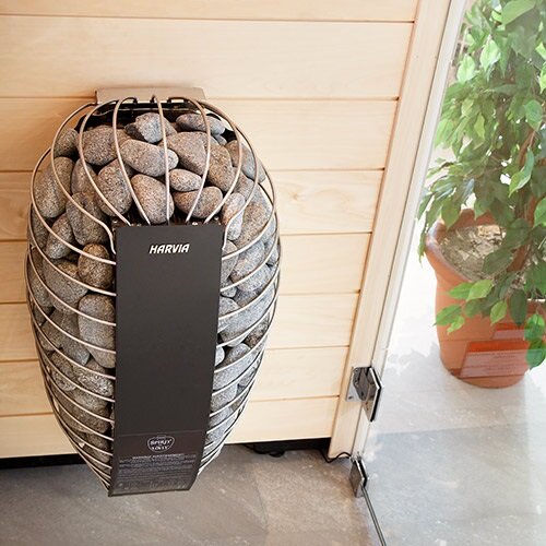 Harvia SP60E electric sauna heater mounted on wooden sauna wall filled with stones near glass door