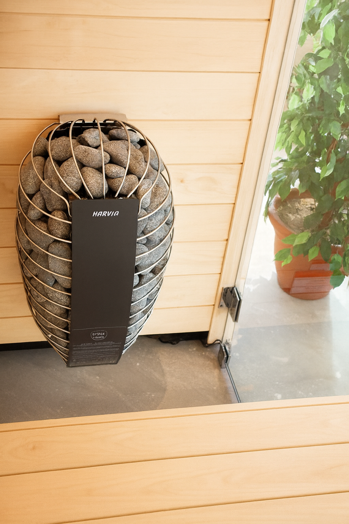Harvia SP80E electric sauna heater mounted in wooden sauna with stacked sauna stones glass door and plant