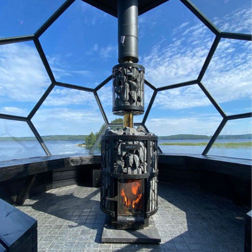 Harvia WHP1500M chimney kit on black wood stove with stone baskets and visible flame inside glass sauna dome overlooking lake