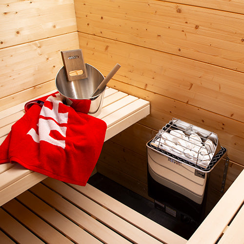 Harvia wall-mounted electric sauna heater beside wooden bench with sauna stones, bucket and red towel