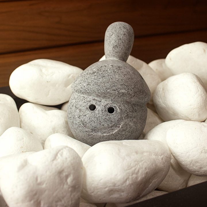 Hukka Elli sauna stone figurine gray smiling pebble accessory surrounded by white sauna stones on wooden background
