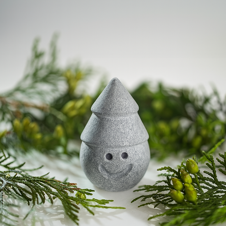 Hukka Jolle Sauna Elf gray stone sauna accessory with smiling face and pointed hat among green spruce branches