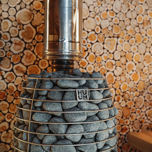 HUUM HIVE Heat Flow Mini 8.5kW sauna heater with piled gray stones in wire cage and stainless chimney against log wall