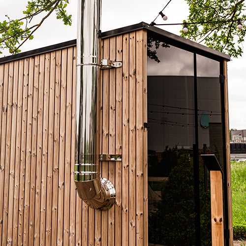 HUUM thru-wall chimney kit installed on modern wooden sauna exterior with metal pipe and glass door