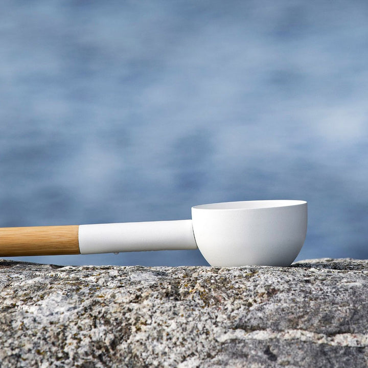 KOLO sauna ladle with wood handle resting on a rock with blue water background