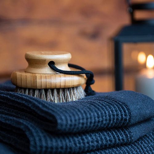 Natural body brush for sauna with wooden handle resting on fluffy towels with a lantern in the background
