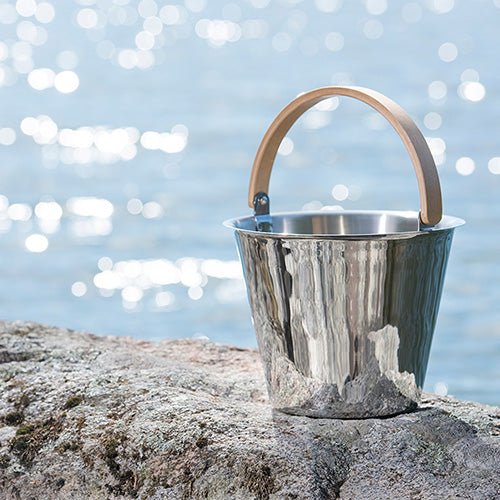 Rento Harri Koskinen sauna bucket in stainless steel with a wooden handle, placed on a rock by the water