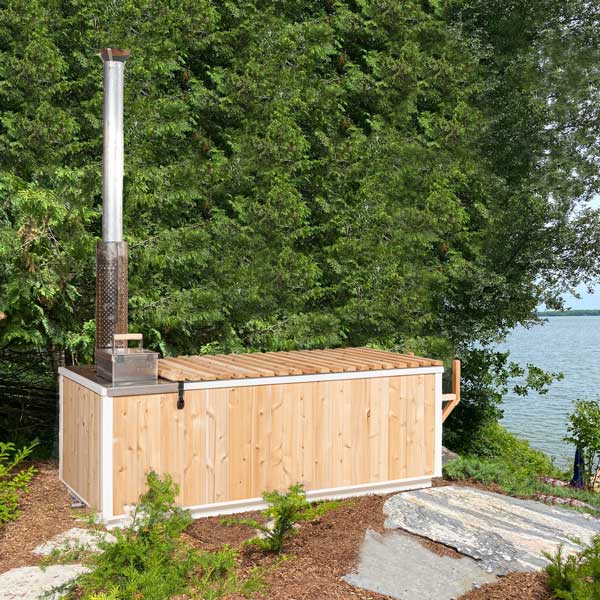 Leisurecraft wood burning hot tub made of light wood panels with metal chimney outdoors near a lake